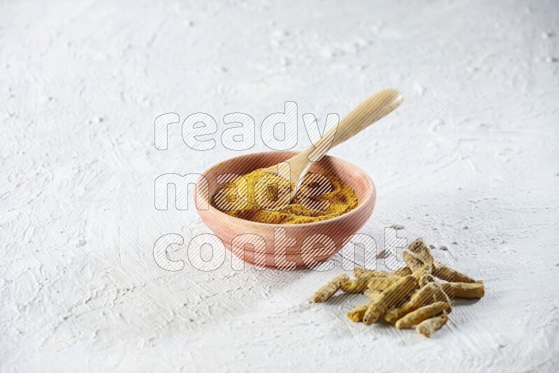A wooden bowl and wooden spoon full of turmeric powder with dried turmeric fingers beside it on textured white flooring