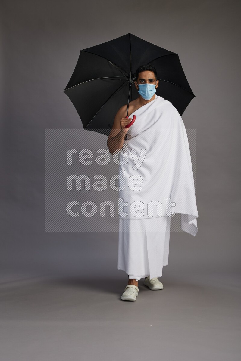 A man wearing Ehram and mask Standing holding umbrella on gray background on gray background