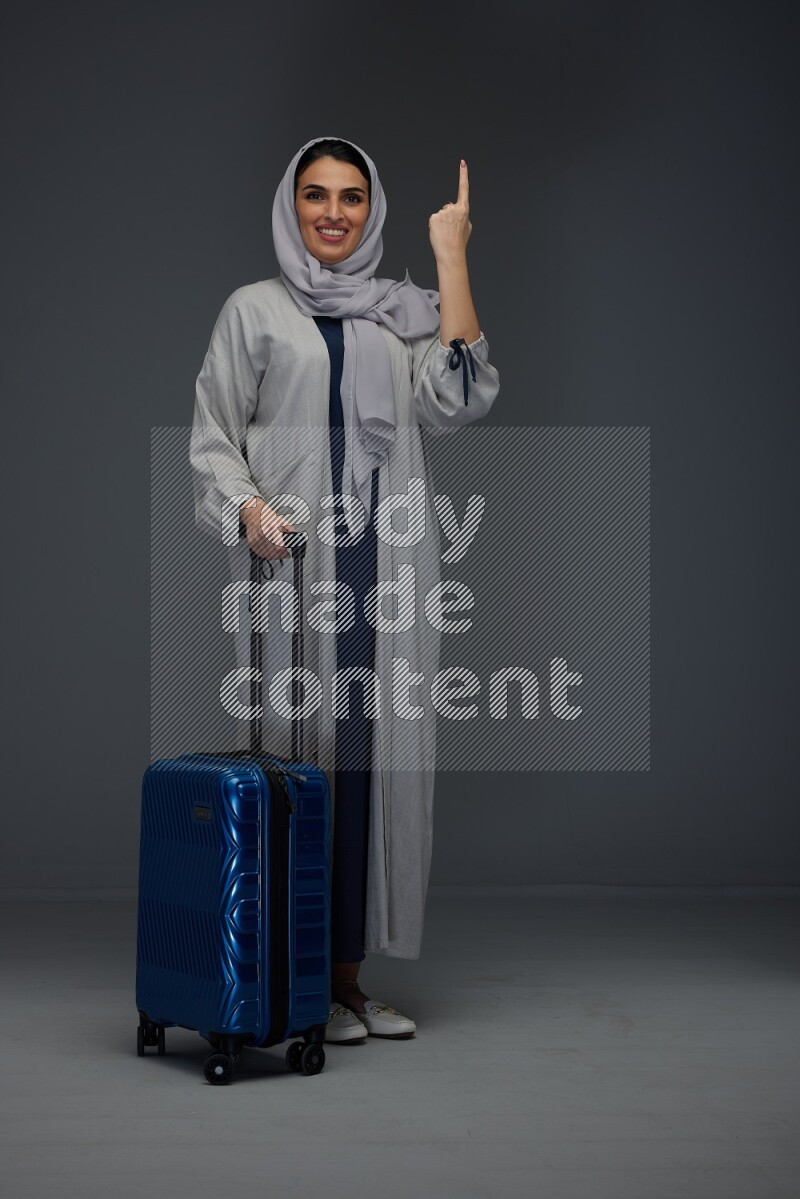 A Saudi woman wearing a light gray Abaya and head scarf standing while holding blue luggage eye level on a grey background