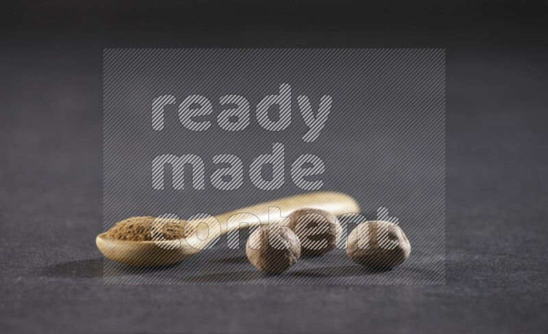 ملعقة خشبية ممتلئة ببودرة جوزة الطيب مع حبوب جوزة الطيب الكاملة بجانبها على خلفية سوداء