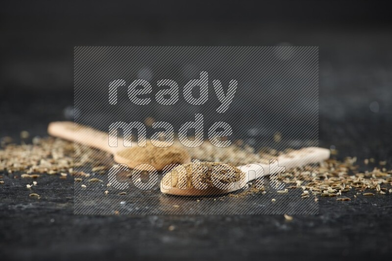 2 wooden spoons full of cumin powder with spreaded seeds on textured black flooring
