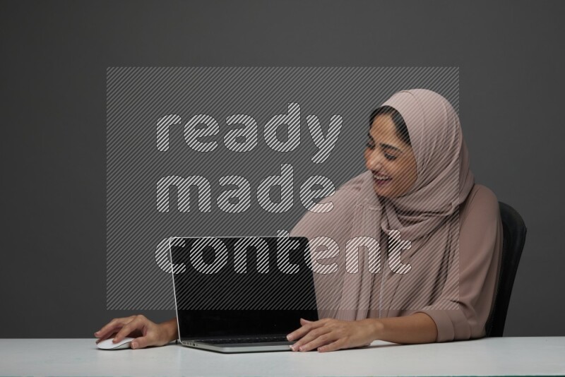 A woman Sitting on her desk  Pointing at her laptop on a Gray Background wearing Brown Abaya with Hijab