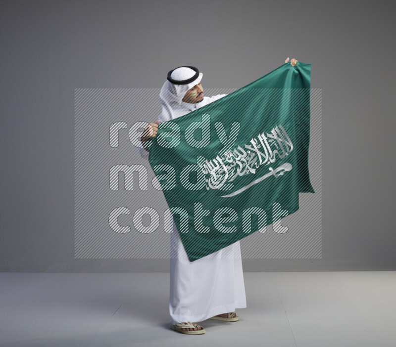 A Saudi man standing wearing thob and white shomag with face painting holding big Saudi flag on gray background