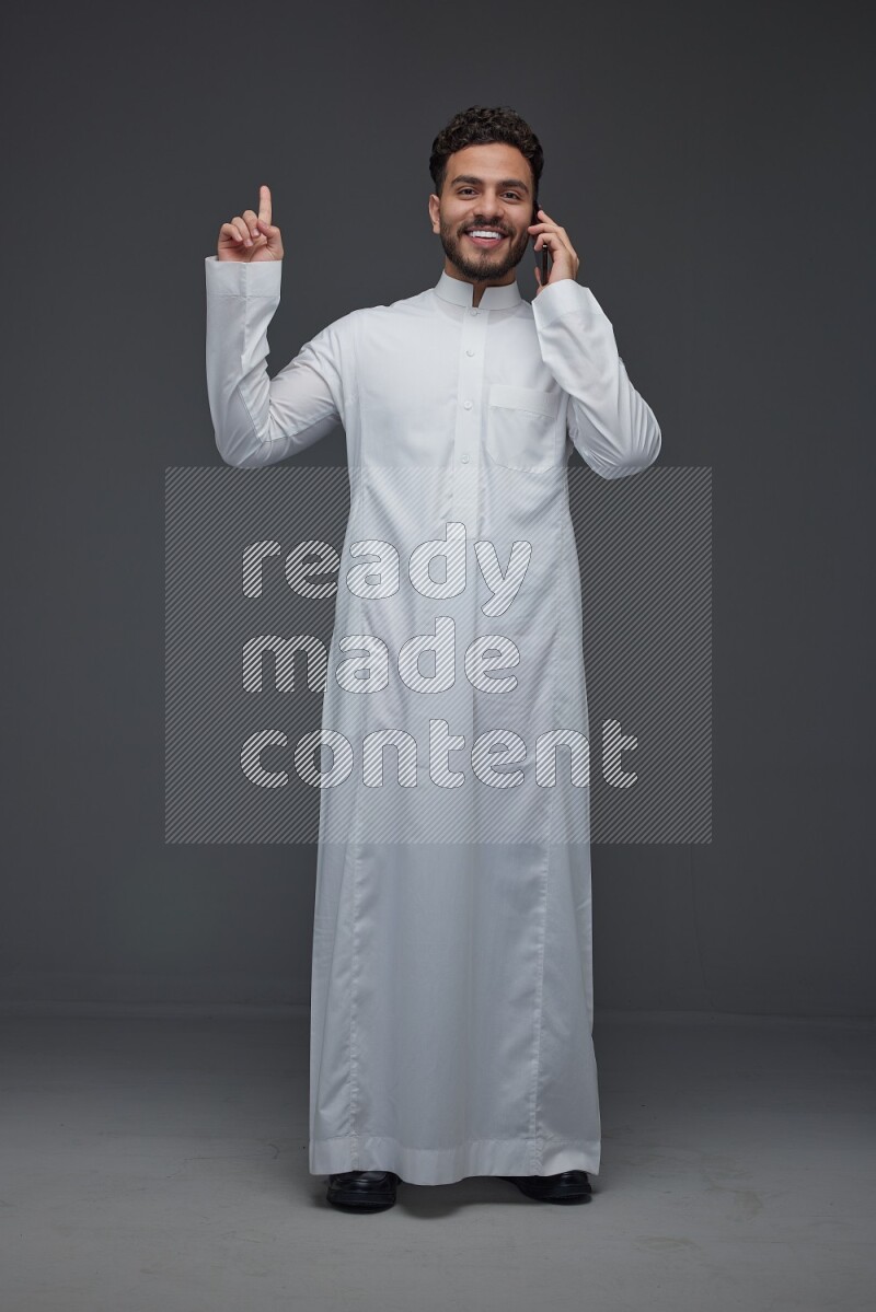 A Saudi man wearing Thobe and talking in the phone while standing and making different hand gestures eye level on a gray background
