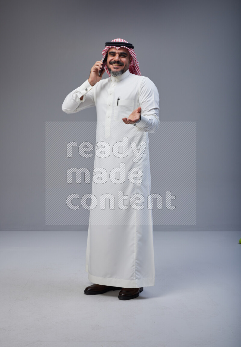 Saudi man Wearing Thob and red Shomag standing talking on phone on Gray background
