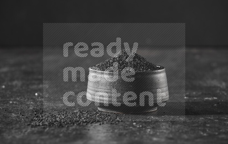 A black pottery bowl full of black seeds on a textured black flooring
