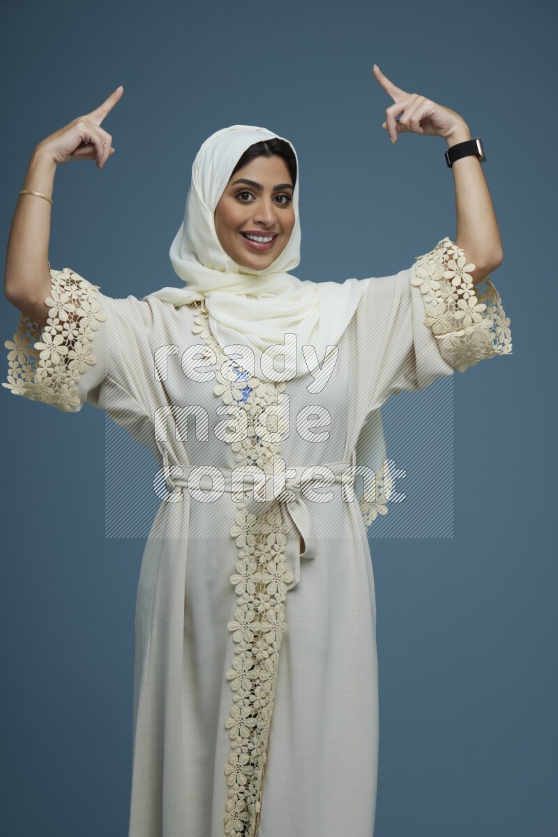 A Saudi woman pointing in a blue background wearing an off-white Abaya Hijab