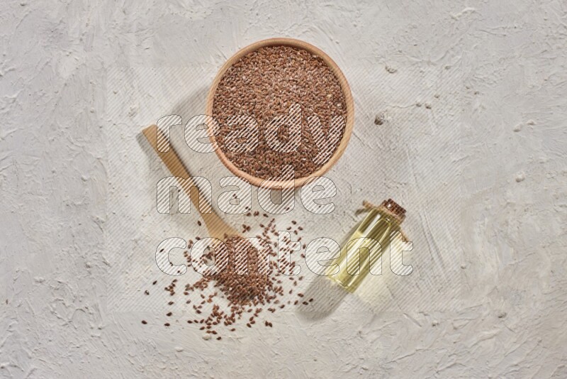 A wooden bowl and spoon full of flaxseeds with a bottle of flaxseeds oil on a textured white flooring
