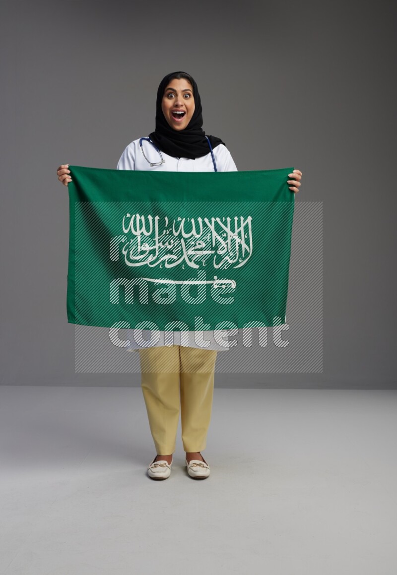 Saudi woman wearing lab coat with stethoscope standing holding Saudi flag on Gray background