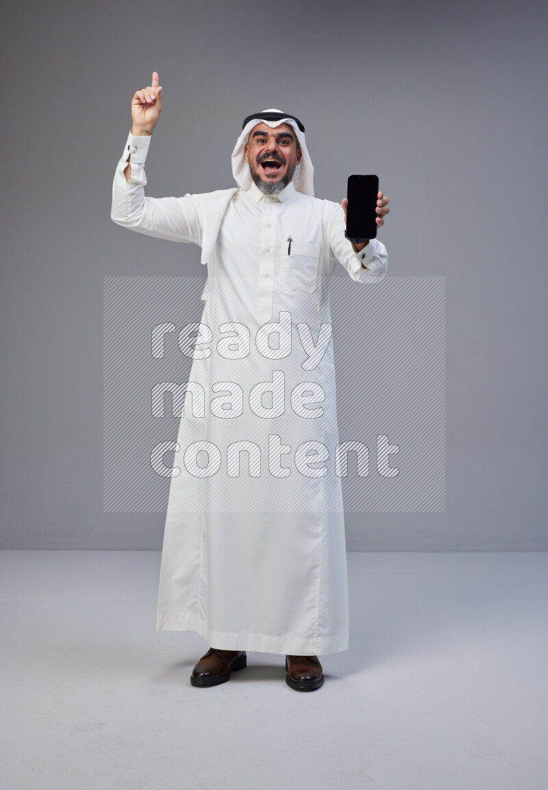 Saudi man Wearing Thob and white Shomag standing showing phone to camera on Gray background