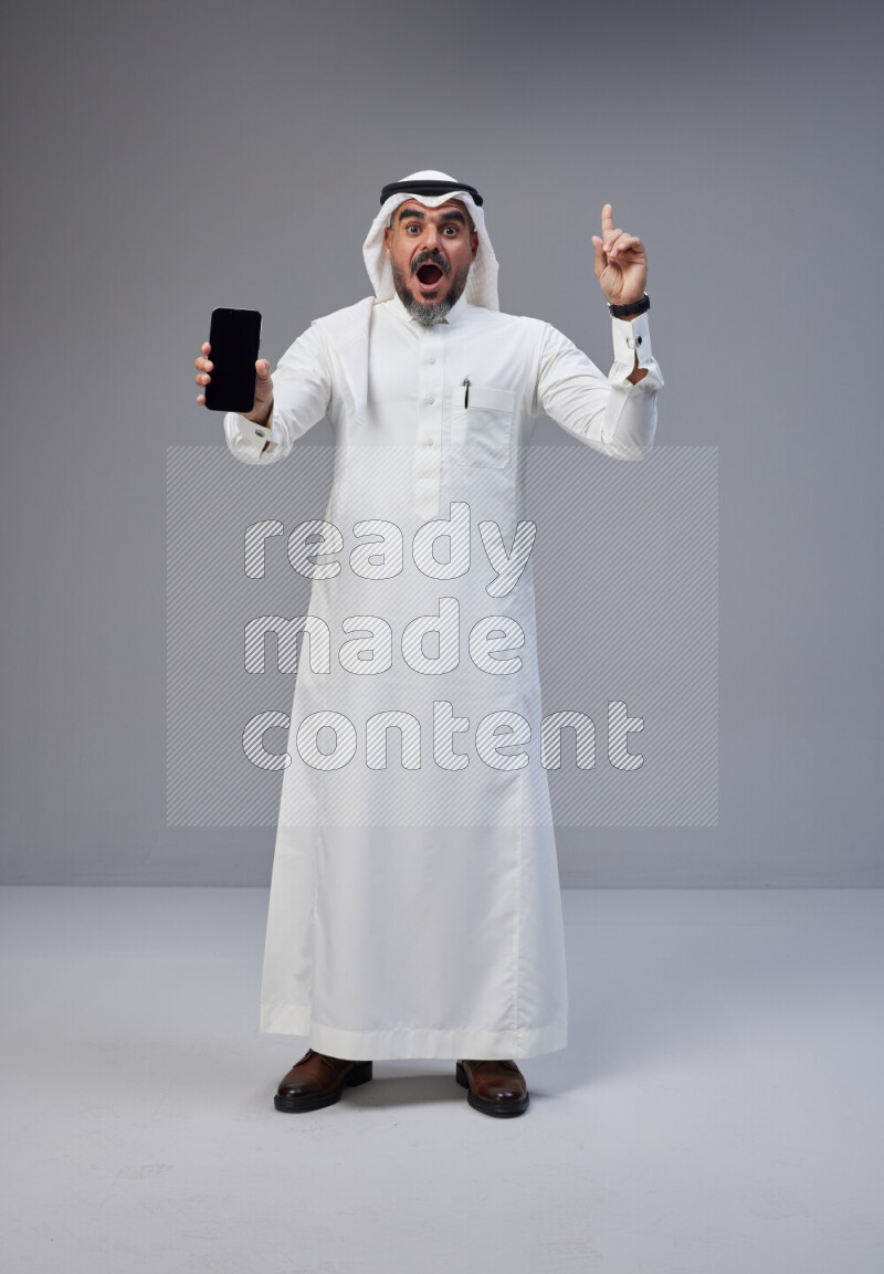 Saudi man Wearing Thob and white Shomag standing showing phone to camera on Gray background