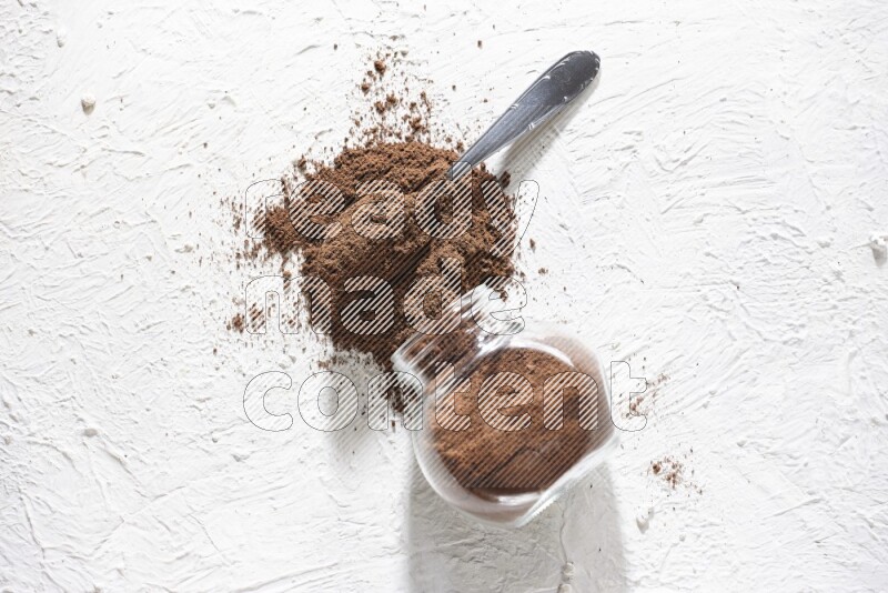 A flipped glass spice jar and a metal spoon full of cloves powder and powder came out of the jar on textured white flooring