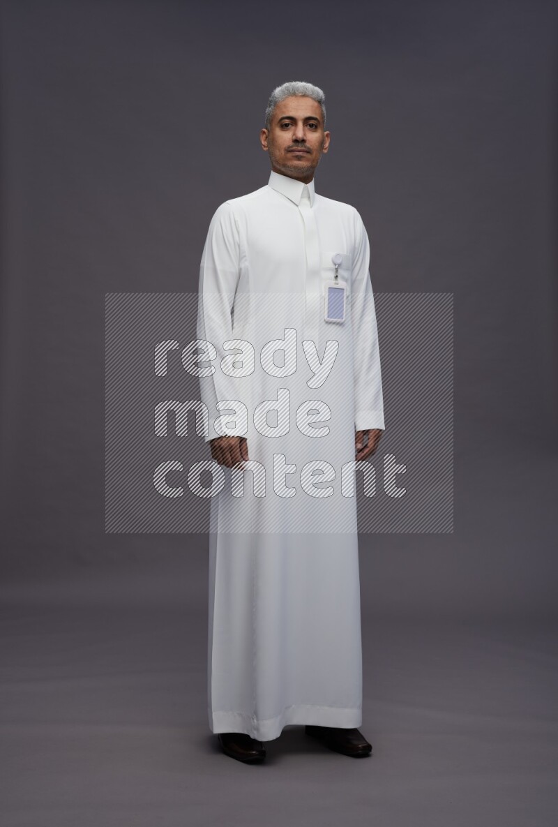 Saudi man wearing thob with pocket employee badge standing interacting with the camera on gray background