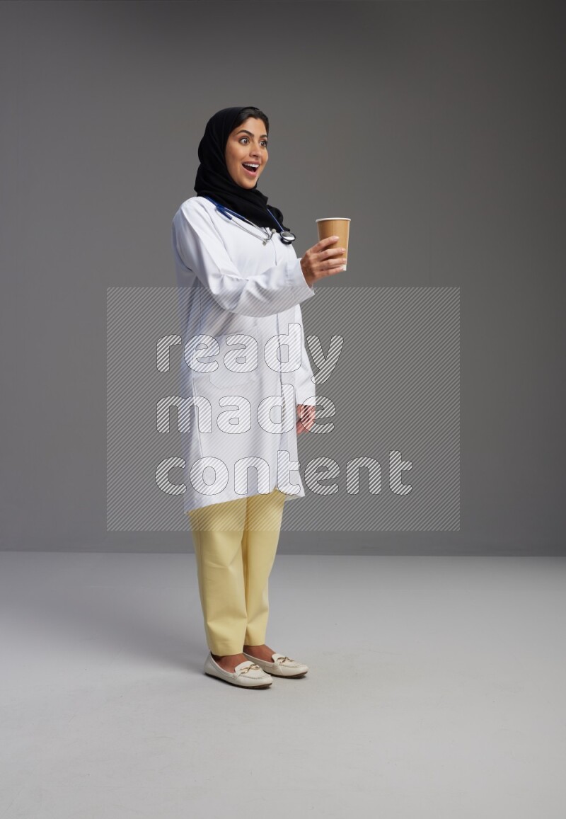 Saudi woman wearing lab coat with stethoscope standing holding paper cup on Gray background