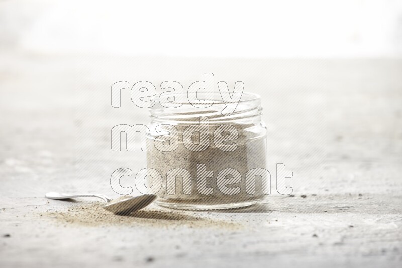 A glass jar and a metal spoon full of white pepper powder on textured white flooring