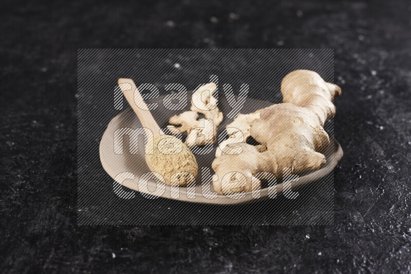 A wooden spoon full of ground ginger powder with fresh and dried ginger, all on a pottery plate on black background