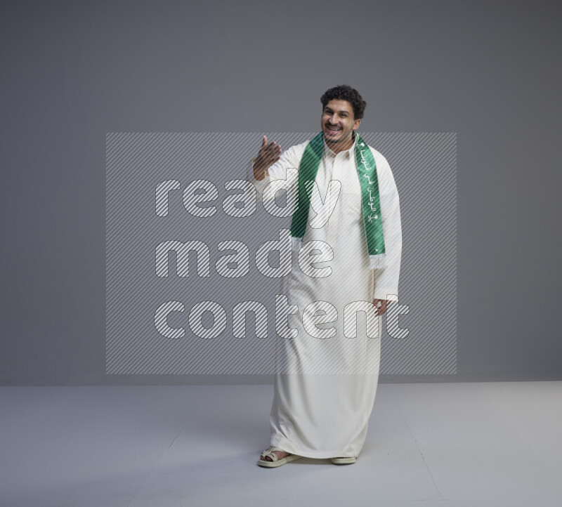 A Saudi man standing wearing thob and Saudi flag scarf on gray background