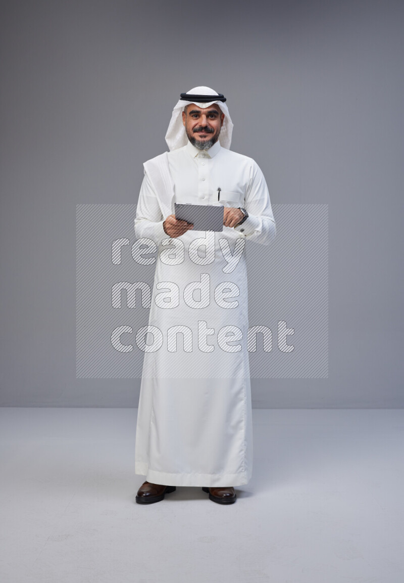 Saudi man Wearing Thob and white Shomag standing working on tablet on Gray background