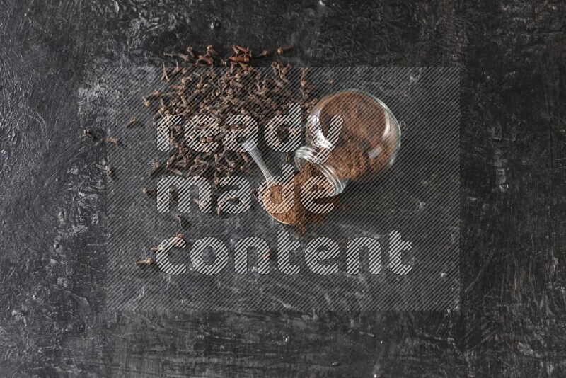 A flipped glass spice jar and a metal spoon full of cloves powder with cloves spread on textured black flooring