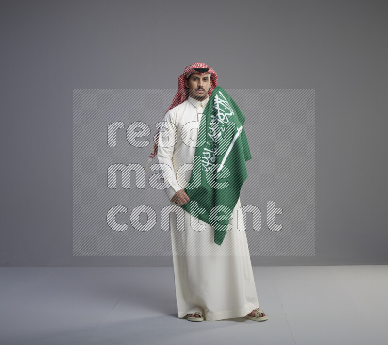 A saudi man standing wearing thob and red shomag holding big saudi flag on gray background