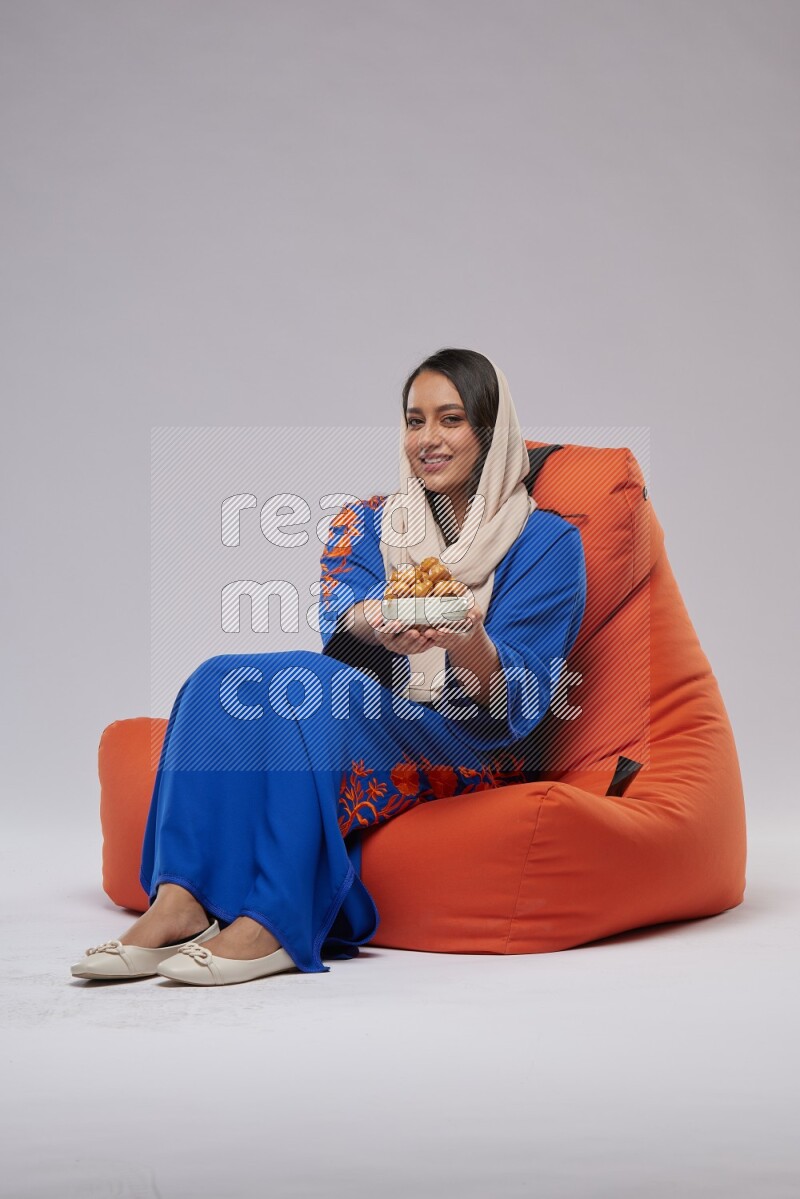 A Woman sitting on an orange beanbag wearing Jalabeya holding a plate of luqaimat