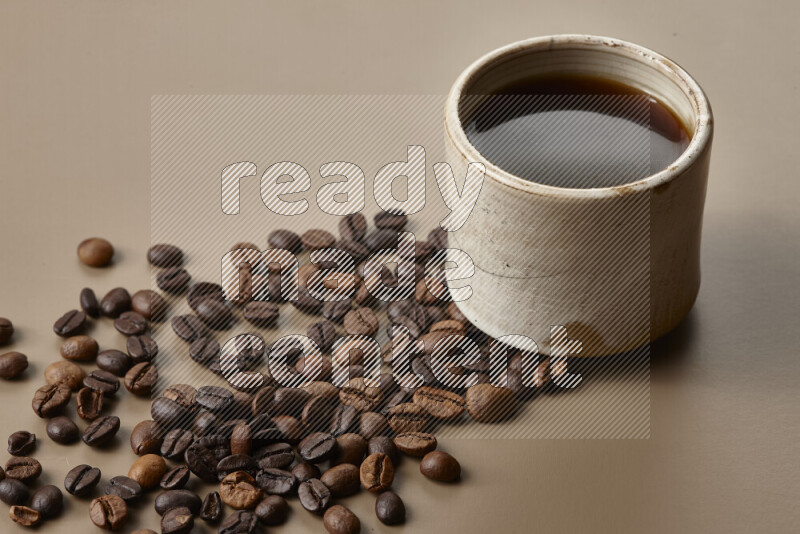 A beige pottery cup of coffee surrounded by roasted coffee beans on beige background