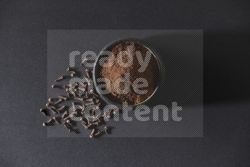 A black pottery bowl full of cloves powder and cloves whole grains on a black flooring