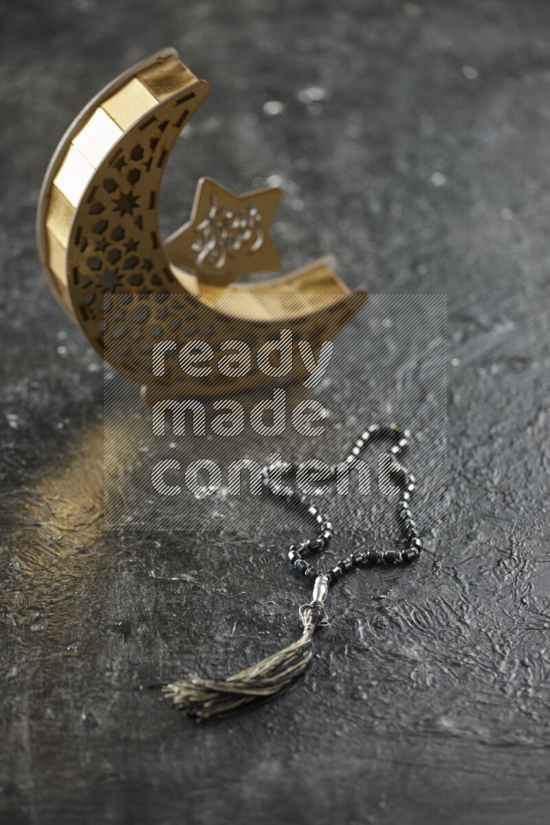 A wooden golden crescent lantern with different drinks, dates, nuts, prayer beads and quran on textured black background