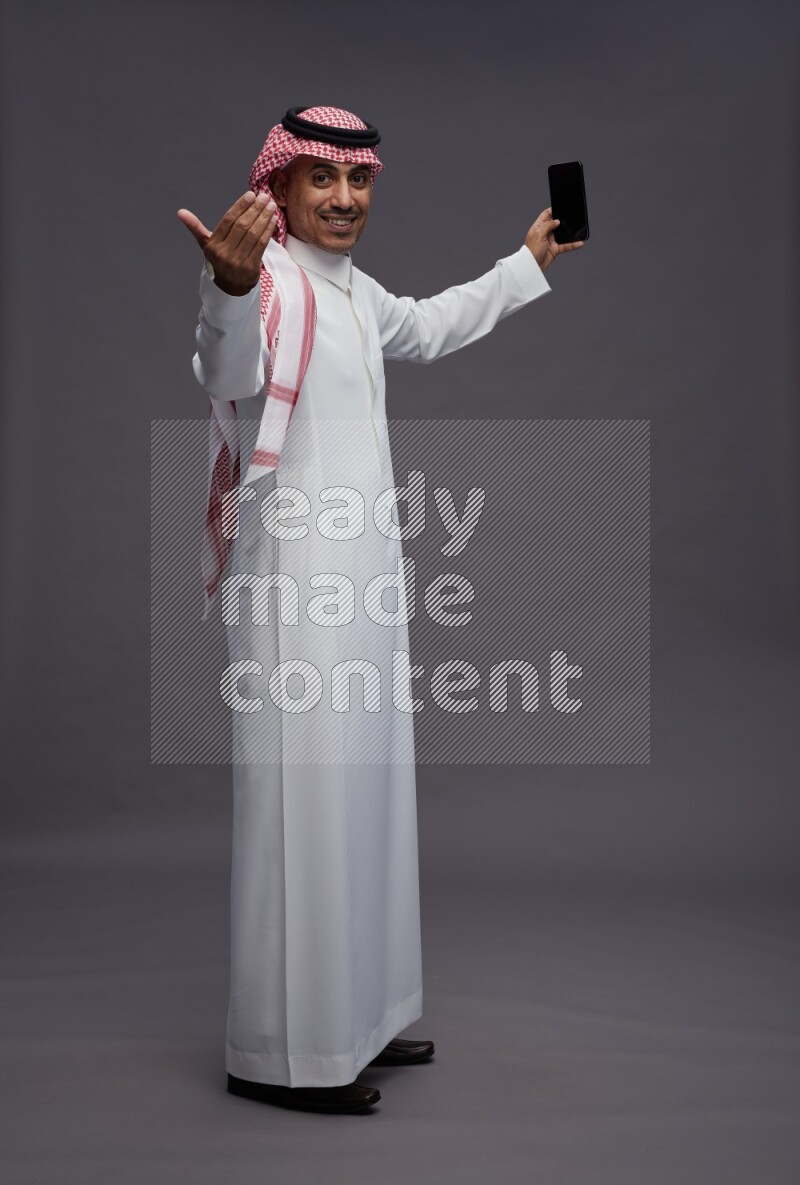 Saudi man wearing thob and shomag standing taking selfie on gray background