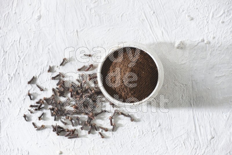 A beige ceramic bowl full of cloves powder and whole cloves on a white flooring