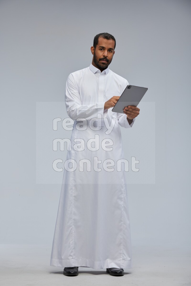 Saudi man wearing Thob standing working on tablet on Gray background