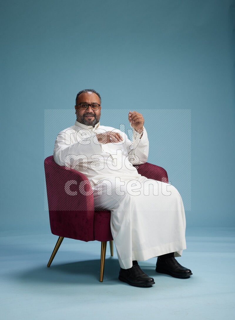 Saudi Man without shimag sitting on chair Interacting with the camera on blue background