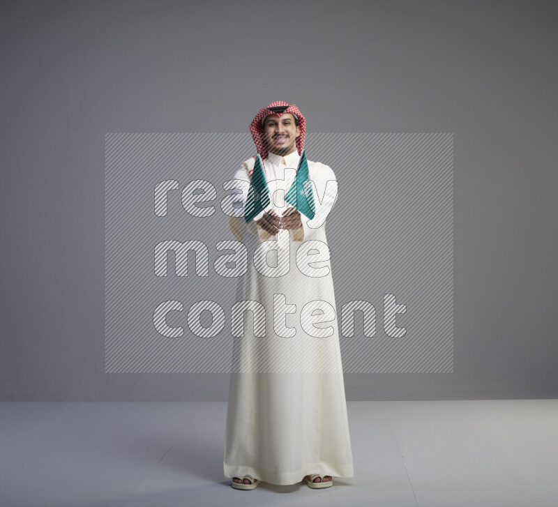 A Saudi man standing wearing thob and red shomag raising small Saudi flag on gray background