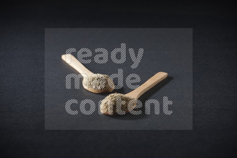 2 Wooden spoons full of cardamom powder on black flooring