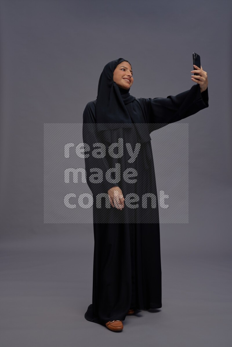 Saudi woman wearing Abaya standing taking selfie on gray background