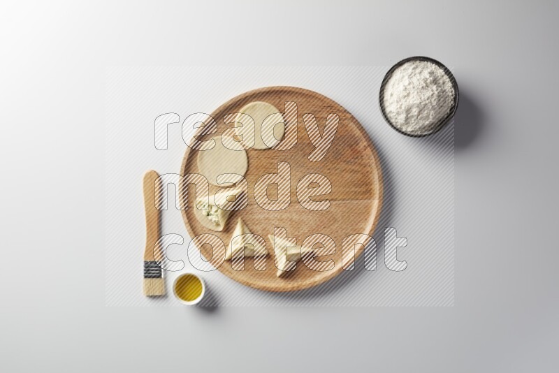 two closed sambosas and one open sambosa filled with cheese while flour, and oil with oil brush aside in a wooden dish on a white background