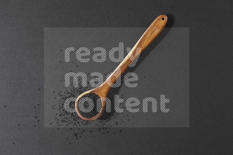 A wooden ladle full of black seeds and the seeds spread beside it on a black flooring