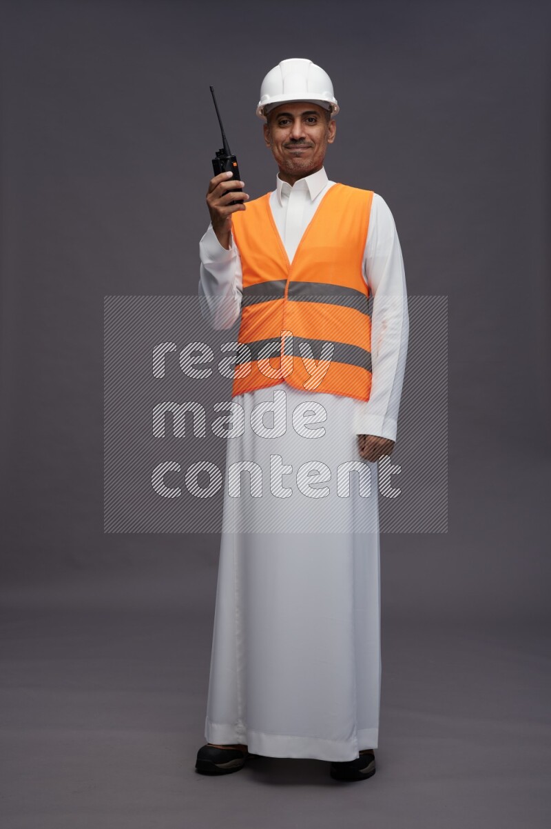 Saudi man wearing thob with engineer vest standing holding walkie-talkie on gray background