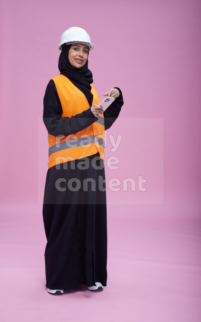 Saudi woman wearing Abaya with engineer vest and helmet standing texting on phone on pink background