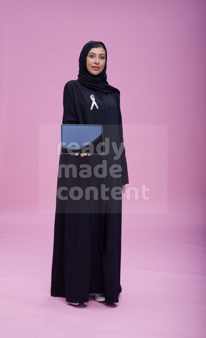 Saudi woman wearing pink ribbon on Abaya standing showing a tablet to camera on pink background