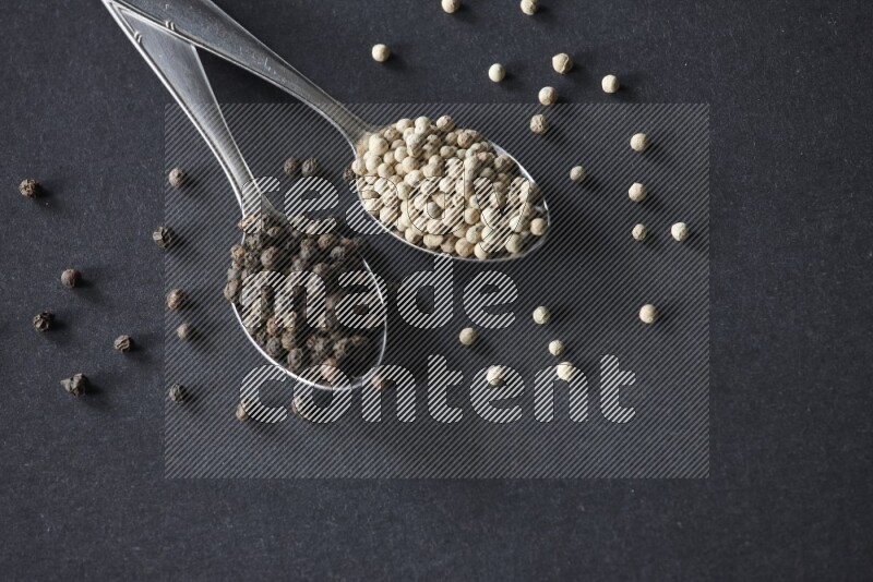 2 metal spoons full of black and white pepper beads with spreaded beads on black flooring