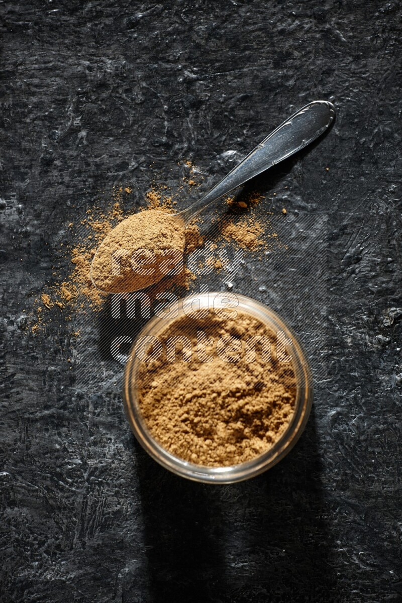 A glass jar and a metal spoon full of allspice powder on a textured black flooring