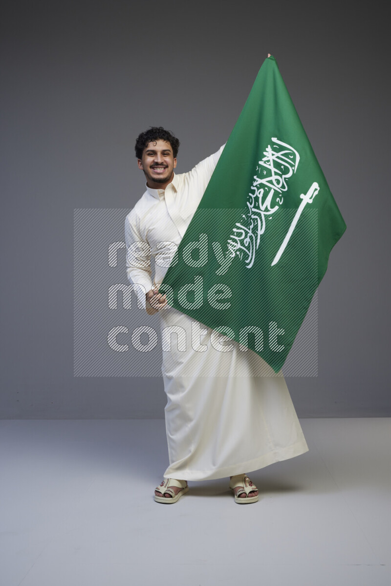 A Saudi man standing wearing thob holding big Saudi flag on gray background