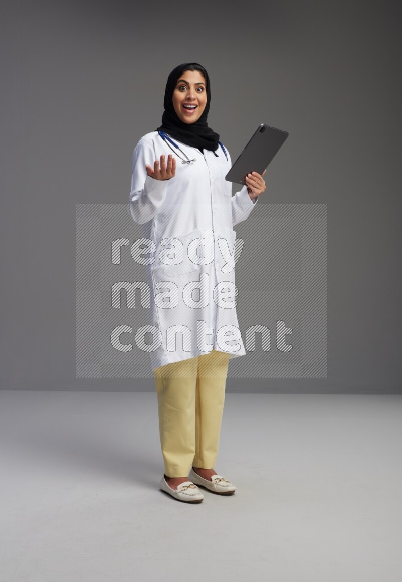 Saudi woman wearing lab coat with stethoscope standing working on tablet on Gray background