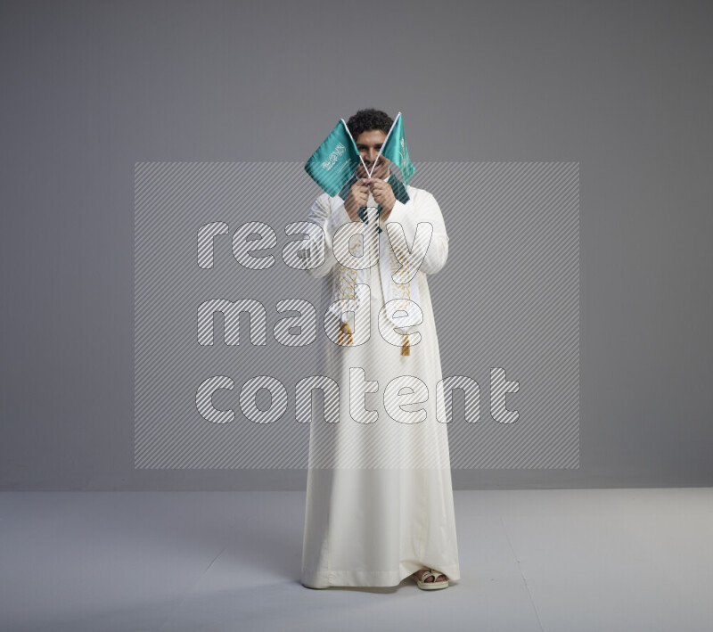 A Saudi man standing wearing thob and saudi flag scarf and holding small saudi flag on gray background