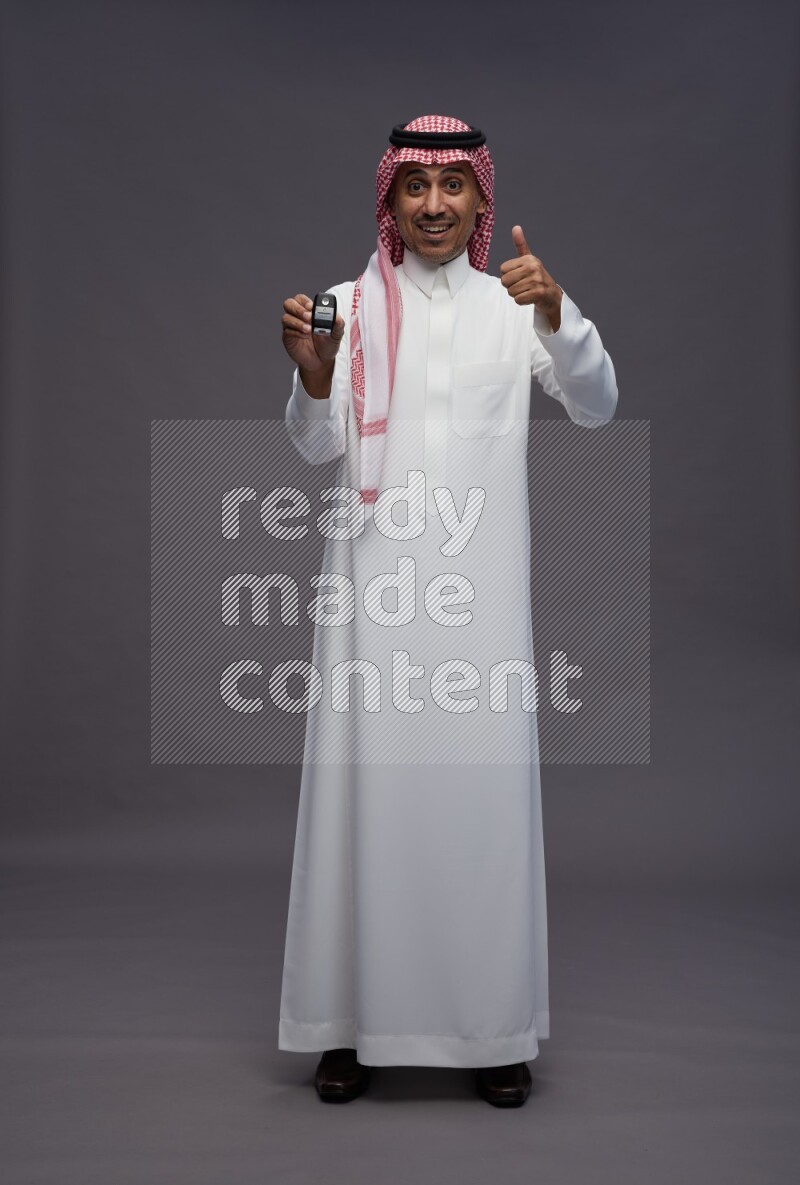 Saudi man wearing thob and shomag standing holding key car on gray background