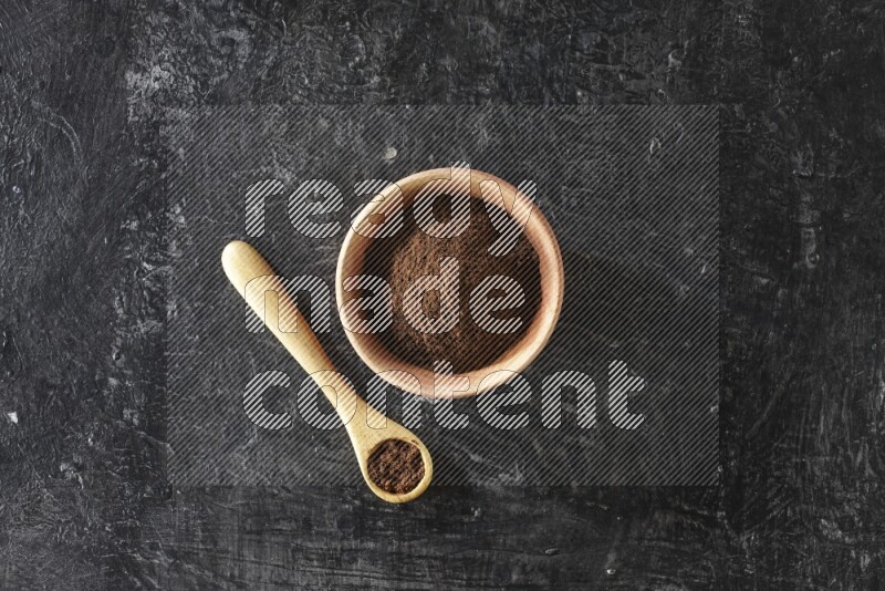 A wooden bowl and wooden spoon full of cloves powder on textured black flooring