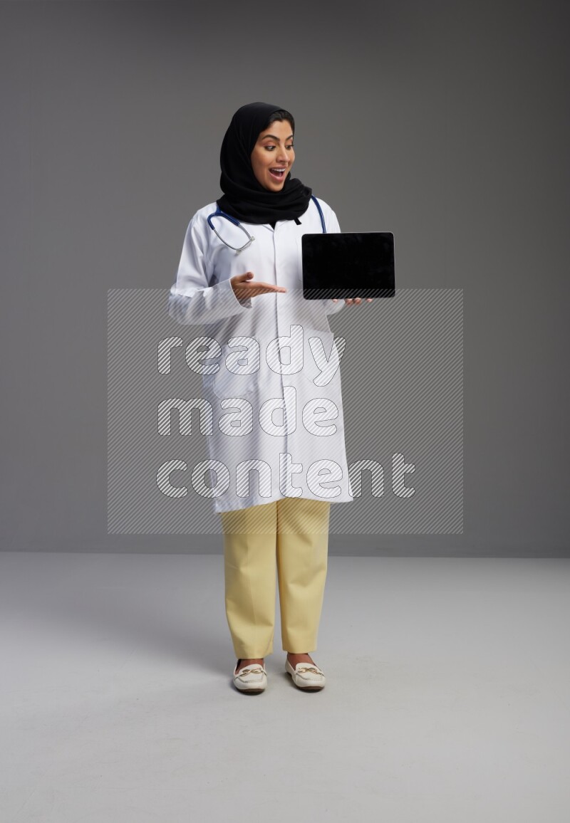 Saudi woman wearing lab coat with stethoscope standing showing tablet to camera with sign in the back on Gray background