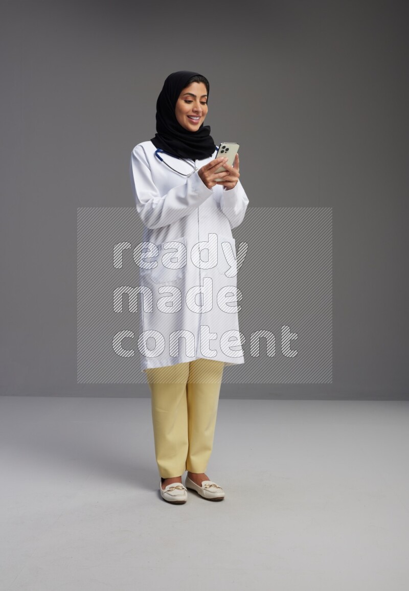 Saudi woman wearing lab coat with stethoscope standing texting on phone on Gray background