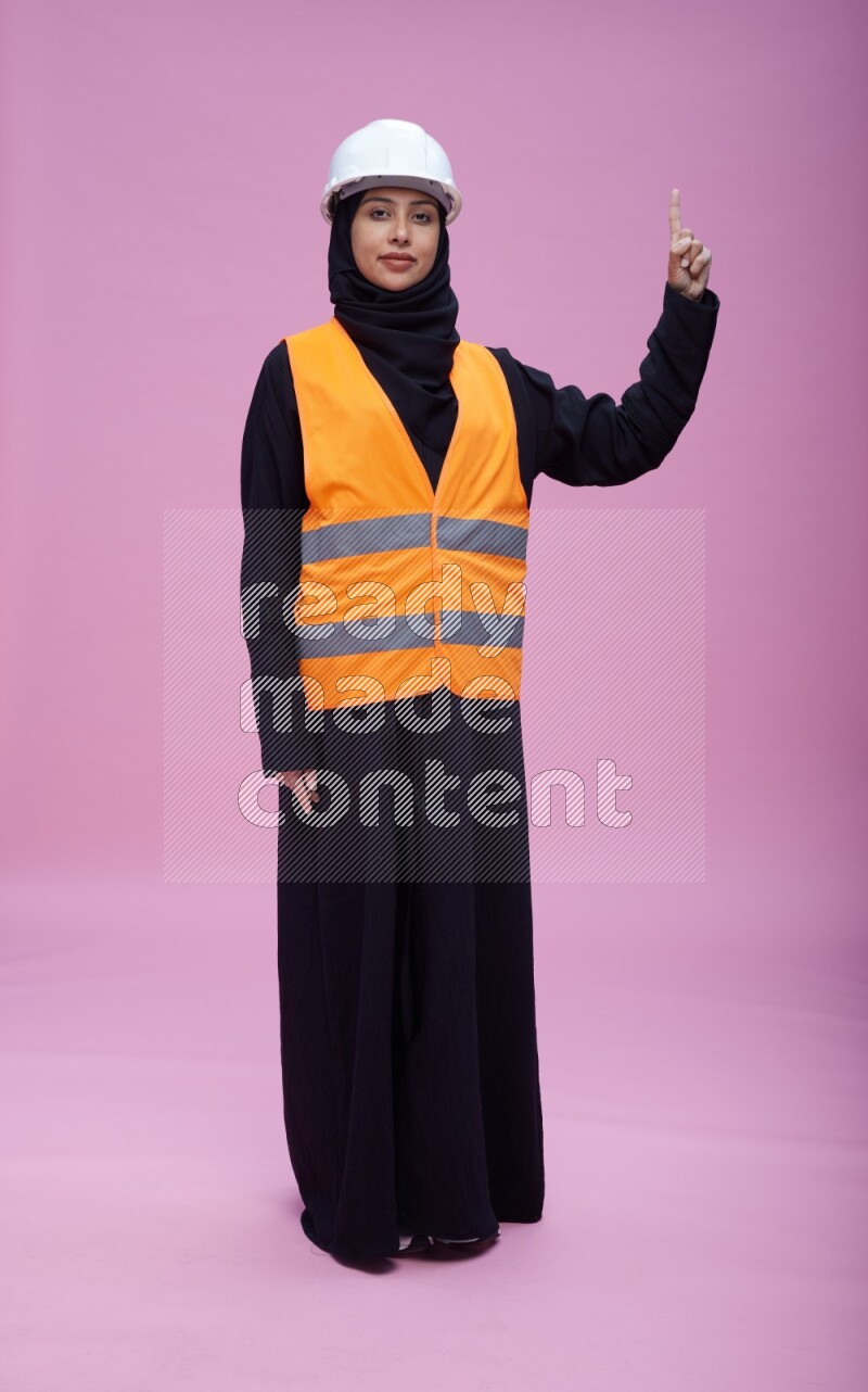 Saudi woman wearing Abaya with engineer vest and helmet standing interacting with the camera on pink background